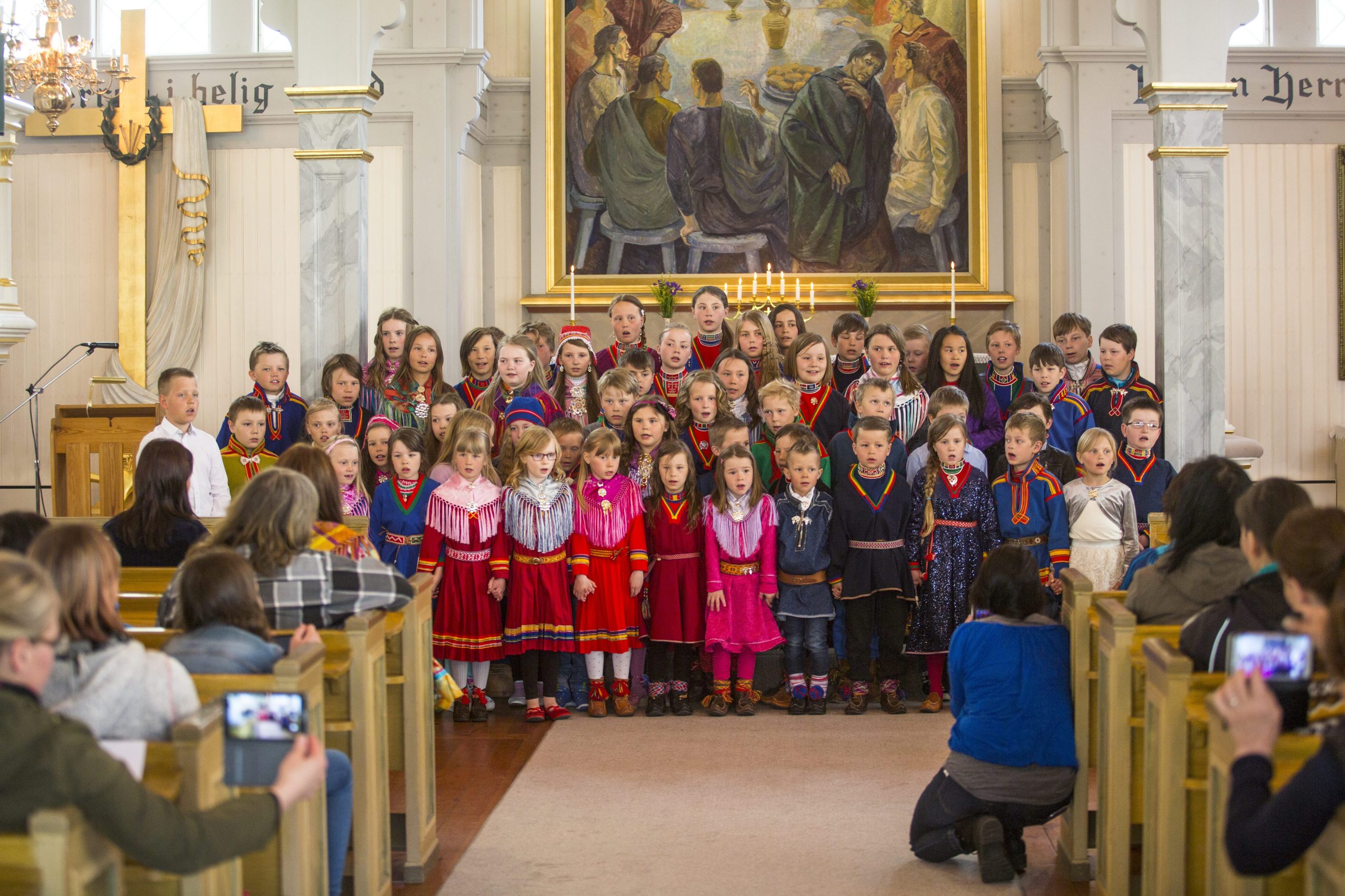 Barn står uppställda för körsång längst fram i en kyrka. Barnen har röda eller blå kläder på sig.
