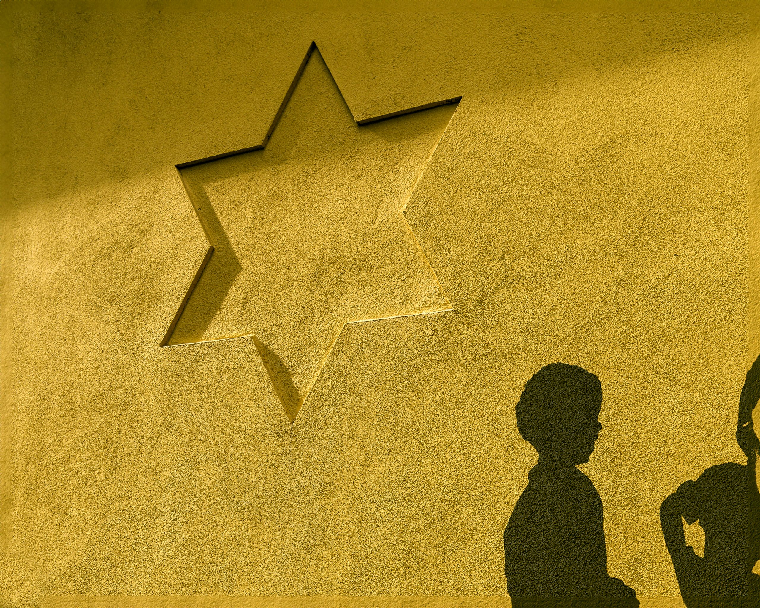 På en gul husfasad syns davidstjärnan i relief. Under stjärnan syns skuggor av människor.