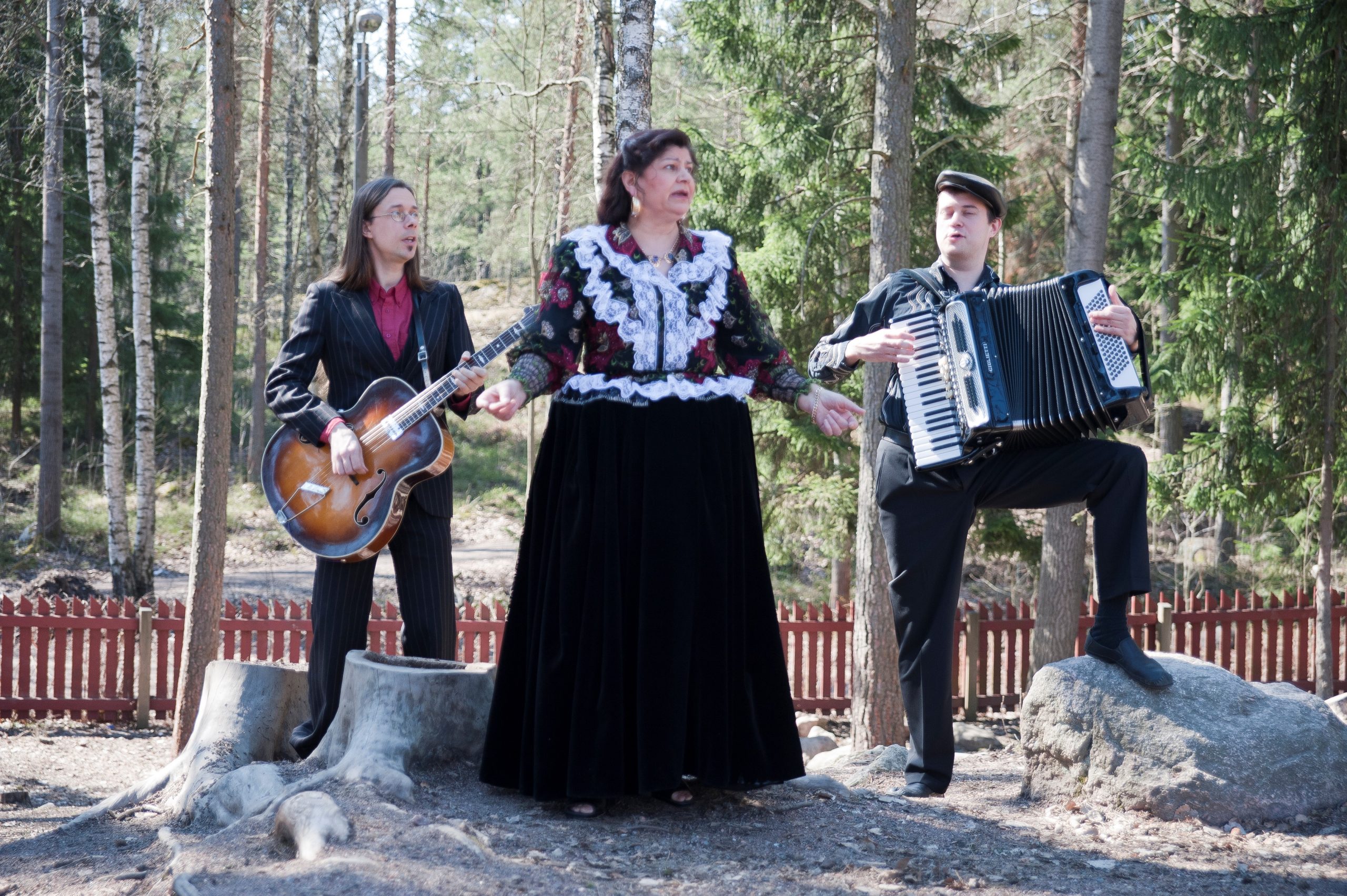 Tre musiker. En gitarrist, en dragspelare och en sångerska. Kvinnan som sjunger har en rikt dekorerad blus och en stor svart kjol.