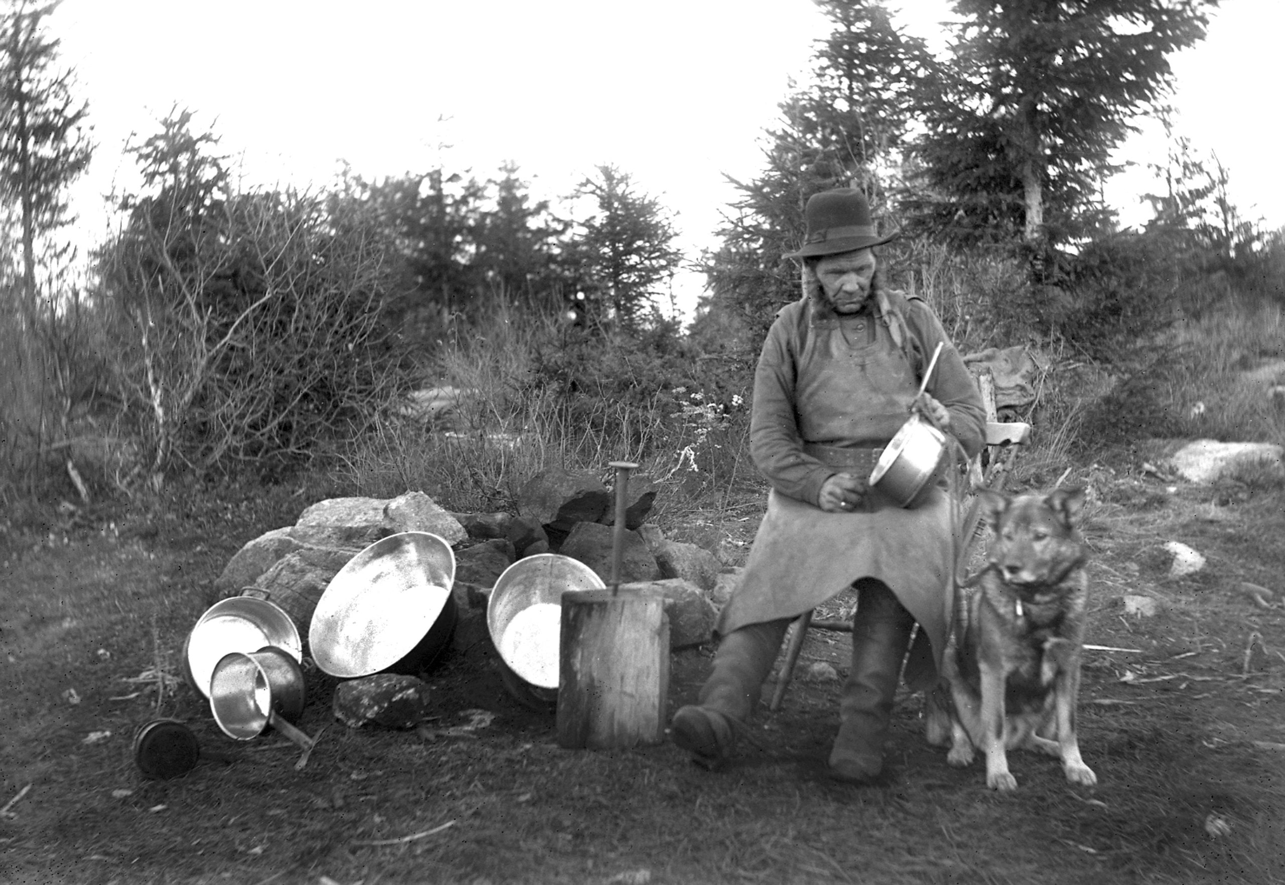 Svartvitt foto av en man i hatt som håller i ett metallkärl. Bredvid honom sitter en hund.