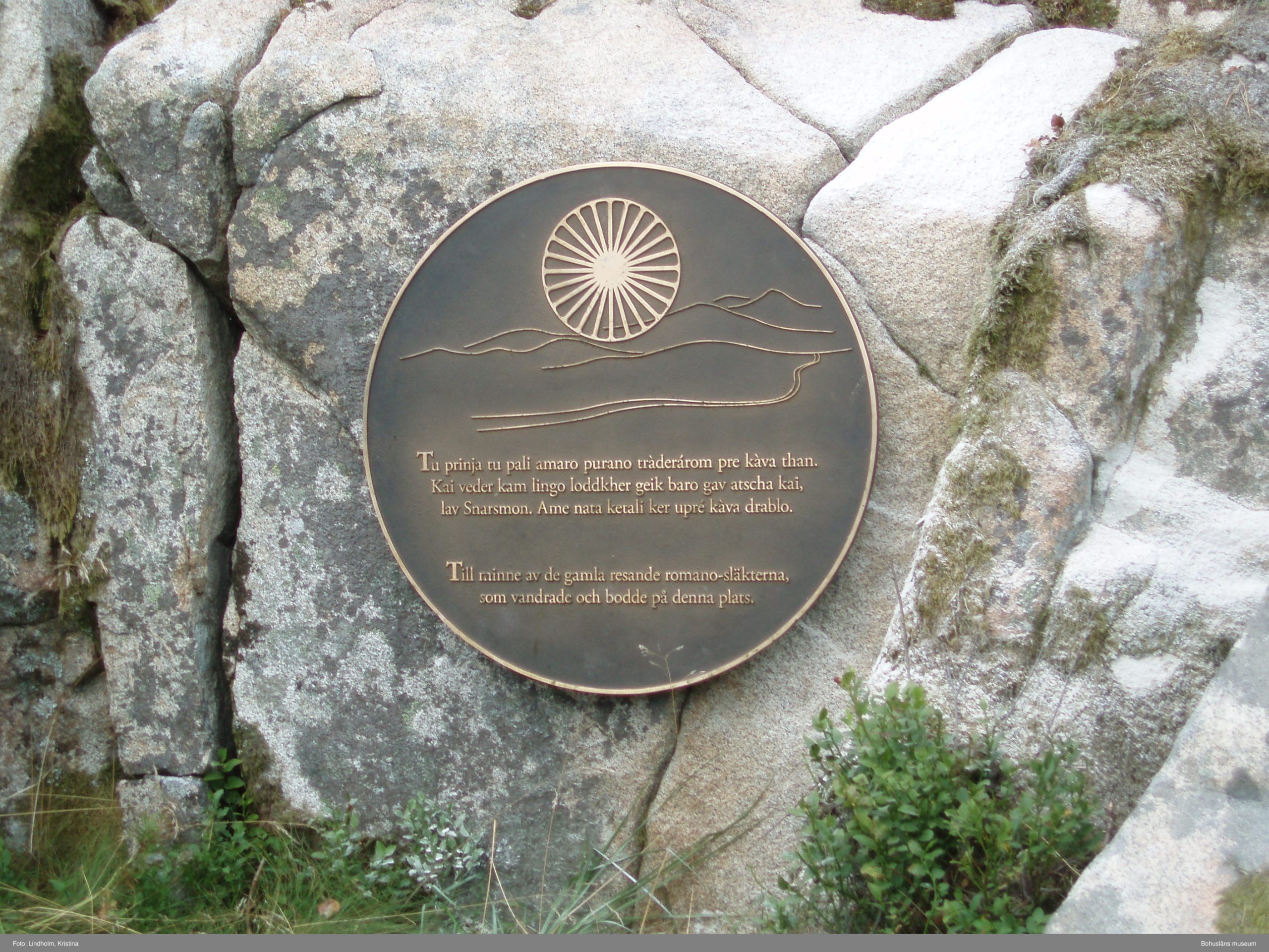 En rund metallskylt monterad på en sten. På skylten syns vagnshjulet, en romsk symbol samt texten "Till minne av de gamla resande romano-släkterna som vandrade och bodde på denna plats".