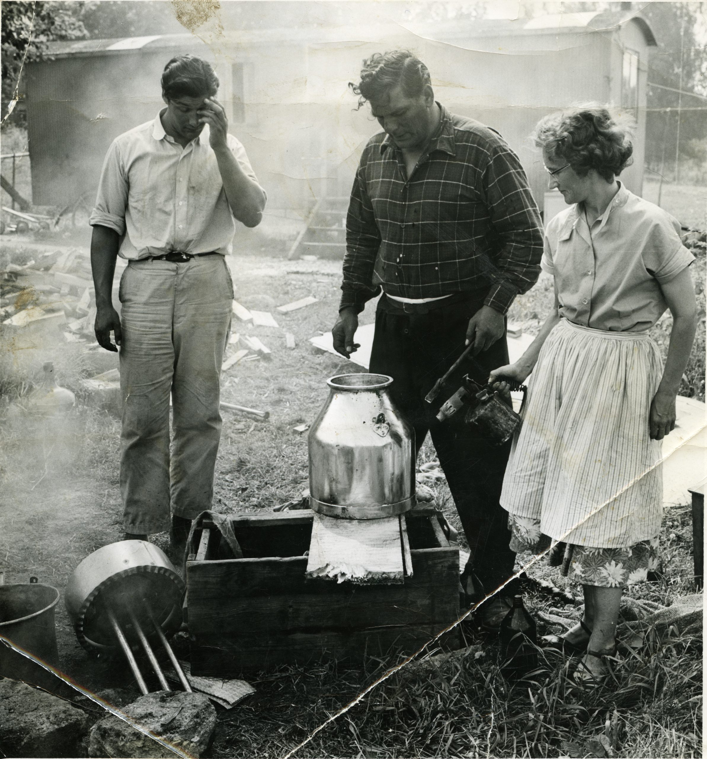 svartvitt foto av två män och en kvinna i medelåldern som står på en gräsmatta och arbetar med ett stort metallkärl. Det är rök omkring.