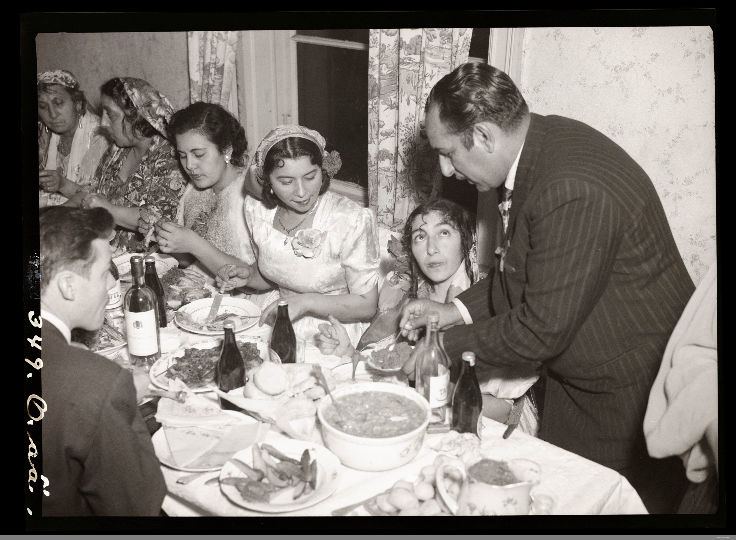 Svartvitt foto av en grupp människor som sitter vid ett långbord under 1950-talet. En mas serverar en av gästerna runt bordet.