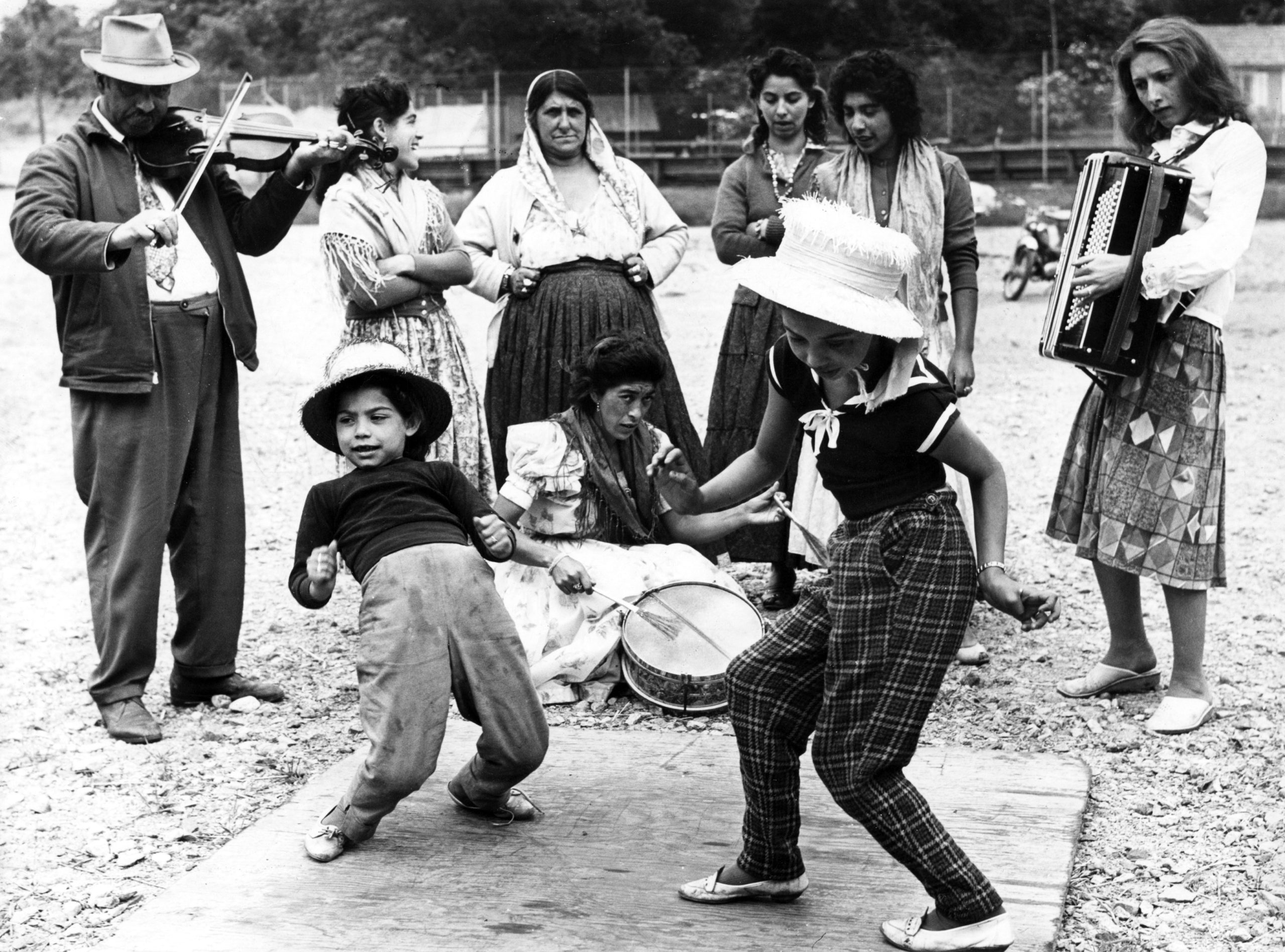 Svartvit foto av en grupp människor. De nuxna spelar instrument och barn dansar til musiken.