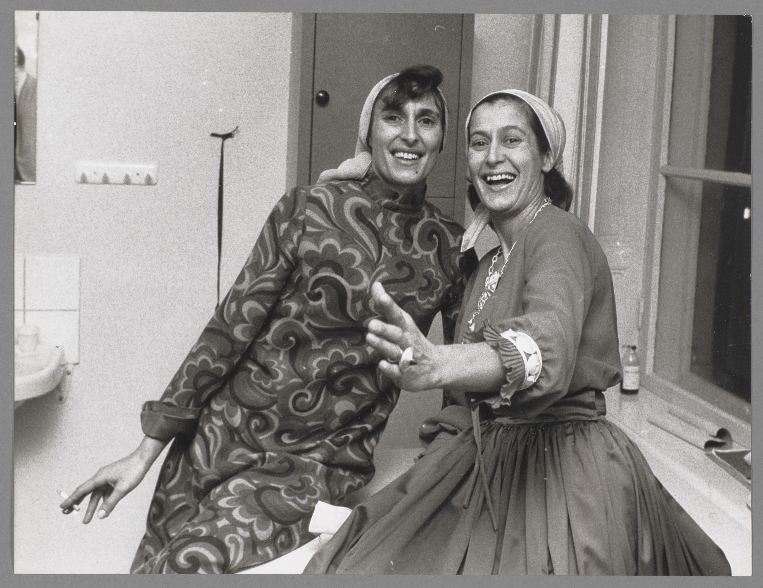 Svartvitt foto av två kvinnor som är fångade i en dansant rörelse. De bär klänningar och sjal på sina huvuden och blickar glatt in i kameran.