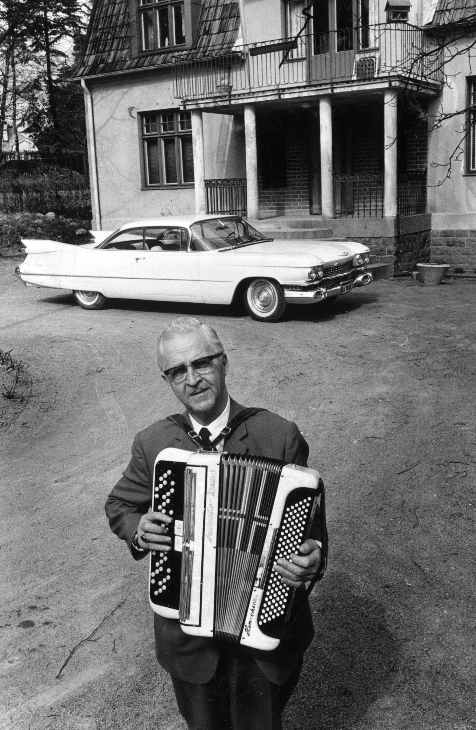 En svartvit bild föreställande en man i 60-årsåldern hållande ett dragspel. Han bär kostym och står på gårdsplanen utanför ett hus, framför vilket en för tiden lyxig amerikansk bil står parkerad.
