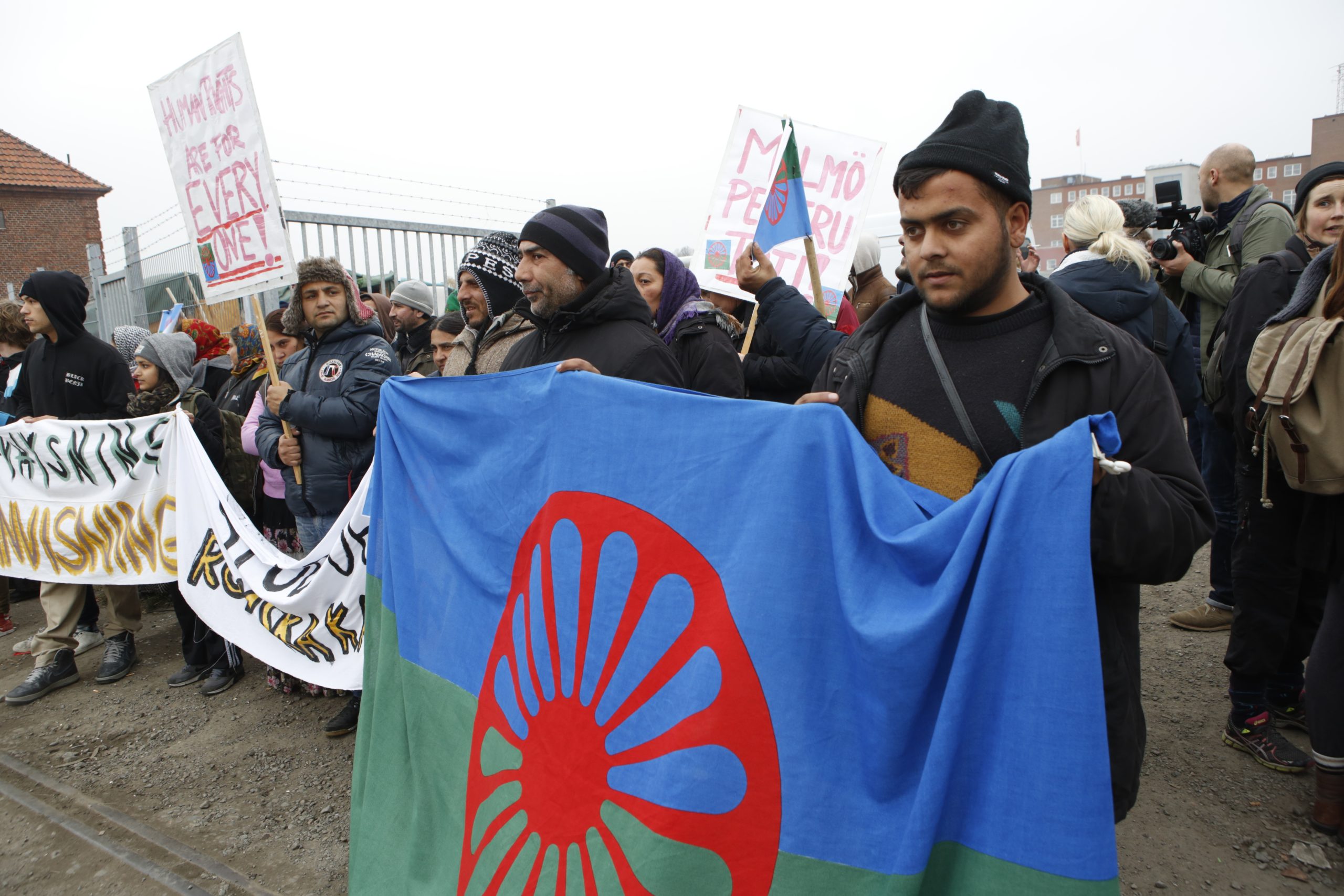 En grupp människor har samlats i vad som ser ut som en demonstration. Några håller upp banderoller och en man håller upp en romsk flagga.