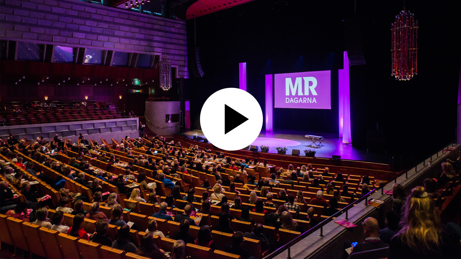 Fotografi av en stor seminariesal med en scen. Ovanpå fotografiet syns en playknapp.