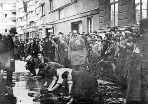 Svartvitt foto av en grupp judiska men tvingas skrubba en gata. Runtomkring står en större grupp människor och tittar på.