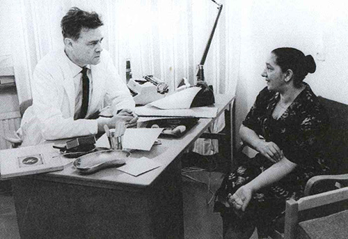 Svartvitt foto av en man i läkarrock som över ett skrivbord pratar med en kvinna i civila kläder.