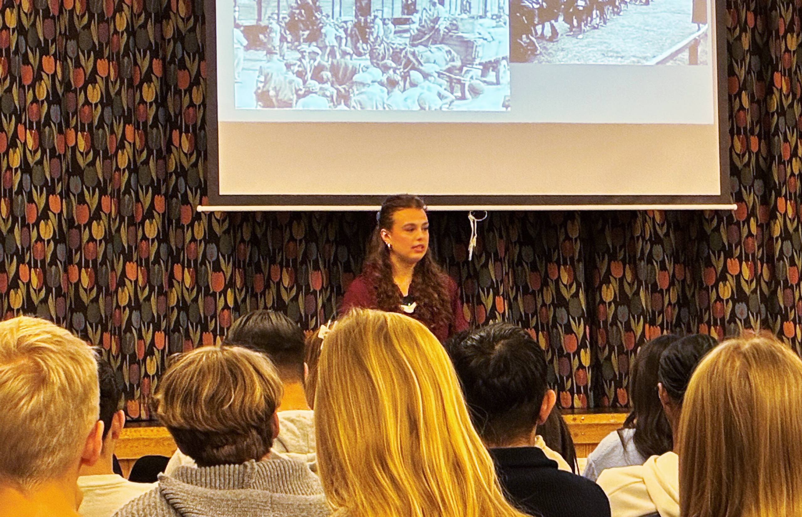 En ung person talar inför en grupp ungdomar. På en projektorduk syns svartvita historiska bilder..